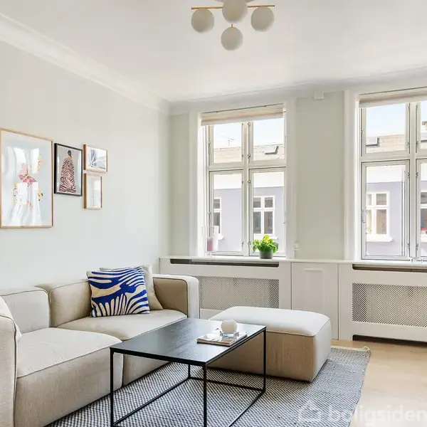 Sofaen står med puder ved et sort bord i en lysegrå stue. På væggen hænger billeder, og store vinduer lader lys ind. En plante pynter i karmen.