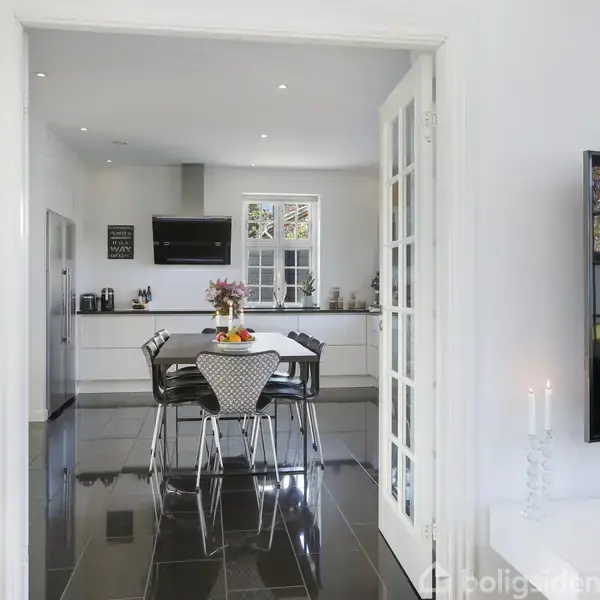 Et stilfuldt køkken vises gennem åbne franske døre. Et bord med stole står på gulvet; blomster og frugter pynter bordet. Rummet har skinnende sorte fliser og hvide vægge.