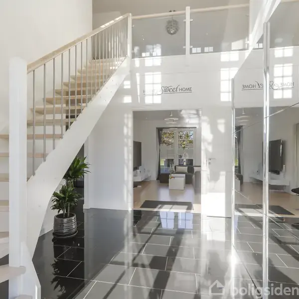 Stilfuld trappe med lyse trin fører opad i en moderne hall med sorte fliser. Indgang til stue ses for enden. Tekst "Home Sweet Home" er skrevet på en væg.