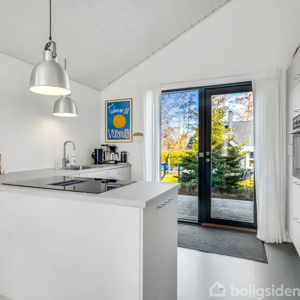 Et moderne køkken med en hvid køkkenø og indbygget kogeplade. Store glasdøre fører ud til en terrasse med udsigt til en have. Væggen har en plakat med teksten "Cachou Lajaunie".