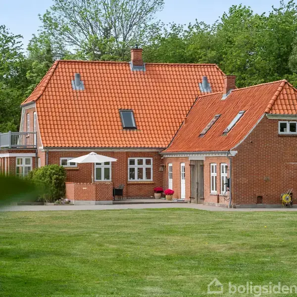 Et rødt murstenshus med rødt tegltag står i en grøn have. Træer omgiver huset, der har flere vinduer og en terrasse.