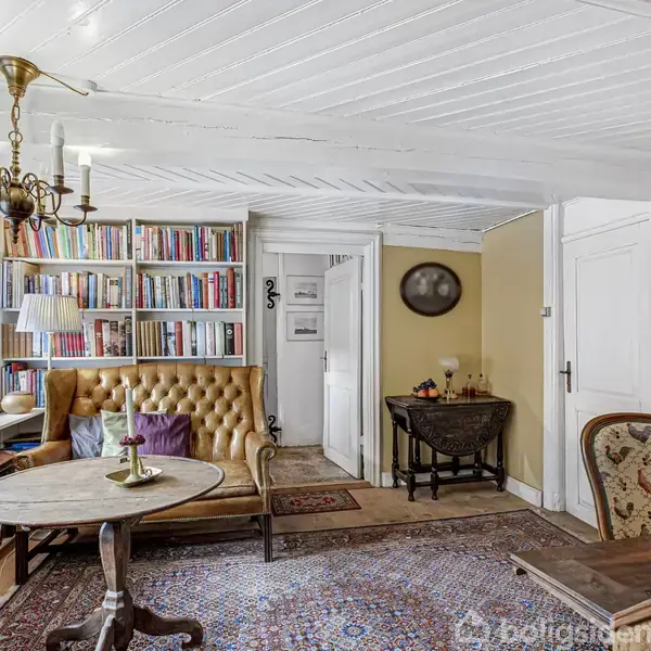 Et hyggeligt stueområde med en brun lædersofa og rundt bord foran en bogreol. En gammel lysekrone hænger fra det hvidmalede loft. Rummet er fyldt med bøger og møbler.