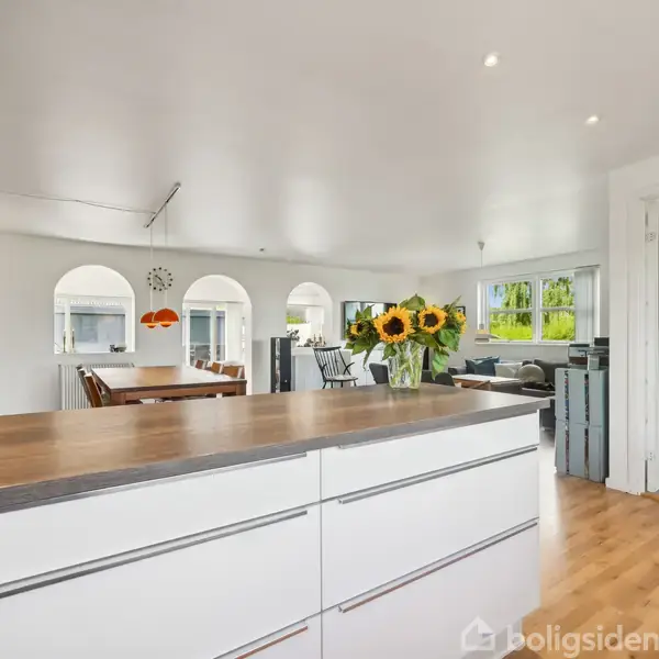 Køkkendisken er dekoreret med en vase af solsikker, der står i et lyst, åbent rum. I baggrunden ses et spisebord med stole og store vinduer.