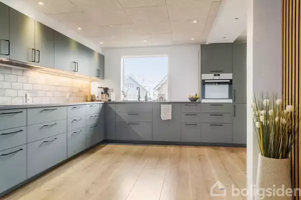 Et moderne køkken med grå skabe. En espressomaskine står på bordpladen, mens en ovn er indbygget i højre væg. Rummet er lyst med trælaminatgulv og hvide vægge.