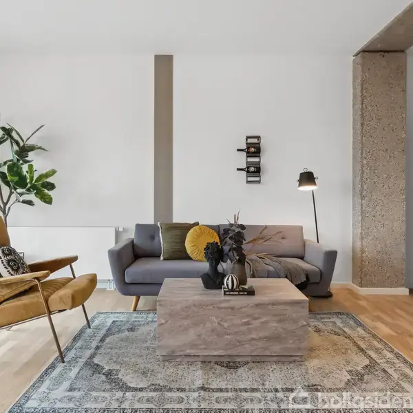 En stue med en grå sofa står centralt, flankeret af en gul lænestol og et stort plantetræ. Et lavt sofabord med dekorationsgenstande står foran sofaen på et mønstret tæppe.