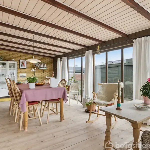 En lys veranda med træloft og -gulv. Et spisebord med en lyserød dug er omgivet af stole. Store vinduer omkranset af hvide gardiner vender ud mod haven.