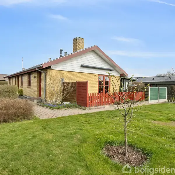 Et enfamiliehus med gul murstensfacade og skråt tag står i en grøn have med græsplæne og buske. Der er en rød træterrasse ved huset og blå himmel i baggrunden.