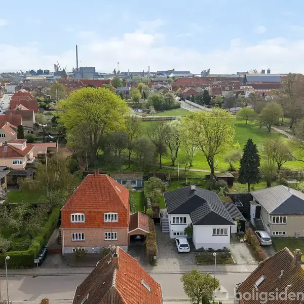 Røde hustage i forgrunden står stille i et boligkvarter, omgivet af grønne træer og parker, med en by i baggrunden under en blå himmel.