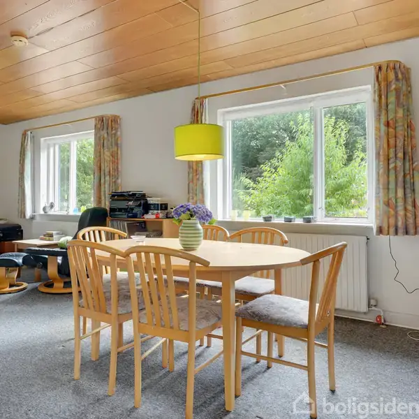 Et spisebord med stole står midt i en stue. En gul lampe hænger over bordet. Stuen har store vinduer med udsigt til grønt, samt en reol og en TV-enhed.
