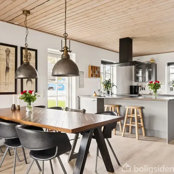 Træspisebord med sorte stole og hængelamper, dekoreret med blomster, i et moderne køkken-alrum med træloft og hvide vægge. Køkkenø med barstole ses i baggrunden.