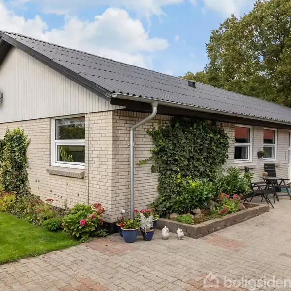 Et murstenshus med sort tag har klatrende planter og blomsterbede langs væggen. Foran er en brolagt terrasse med havemøbler. Græsplæne og træer omkranser huset under en blå skyet himmel.