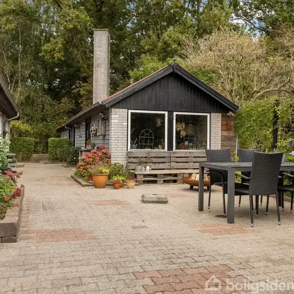 Et lille hus med store vinduer står i en brolagt have med havemøbler og potteplanter. Omgivet af frodig vegetation og træer, giver omgivelsen en rolig atmosfære.