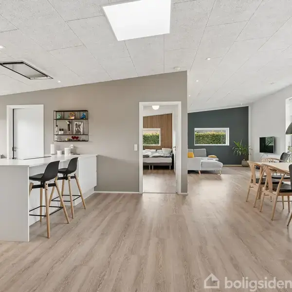 Moderne køkken-alrum med køkkenø, barstole og spisebord med stole. Handling: Køkkenet er åbent med udsigt til en opholdsstue. Kontekst: Lyst trægulv, hvide vægge og store vinduer, naturligt lysindfald.