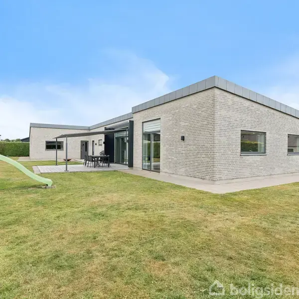 Et moderne enfamiliehus med fladt tag er placeret på en velholdt græsplæne. Ved siden af huset er der en legeplads med en grøn rutsjebane. Himlen er klar og blå.