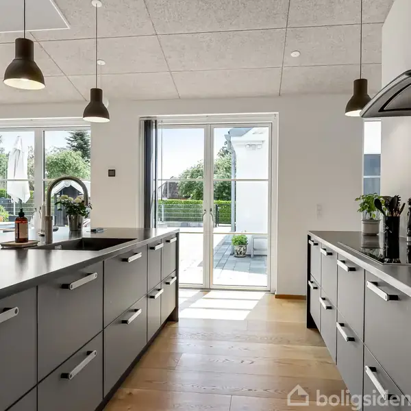 Køkken med sorte bordplader og grå skuffer, står i skarp sollys. Glasdøre fører ud til en terrasse med grøn have. Lamper hænger fra loftet, skaber en moderne atmosfære.