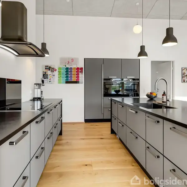 Køkken med grå skabe og sorte bordplader; hængelamper lyser rummet op. Moderne apparater indgår, som indbygget ovn. Trægulv og hvide vægge fuldfører det stilrene design.