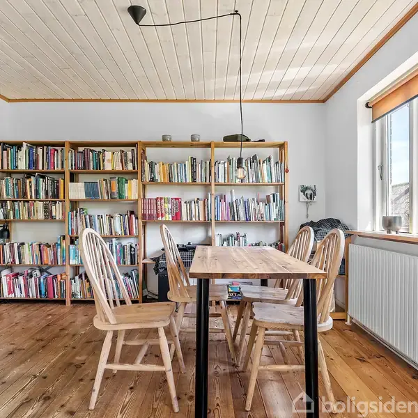 Et spisebord omgivet af træstole står i et lyst rum med trægulv. Hvide vægge har reoler fyldt med bøger. Store vinduer giver udsyn til en grøn have.