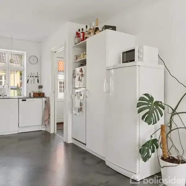 Et hvidt køkken med sorte accenter rummer et køleskab, mikroovn, og planter. Et gennemgående lyst design med vinduer modsat opvaskebordet lader naturligt lys strømme ind.