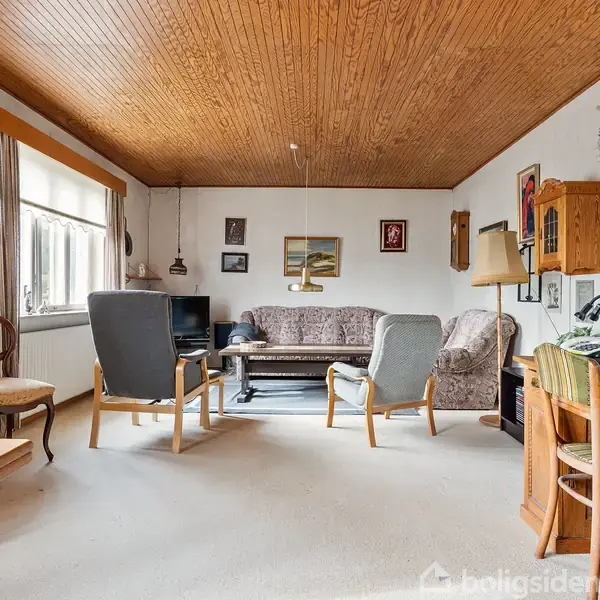 En stue med træloft indeholder en sofa og lænestole, der står omkring et bord. Der er en reol, malerier på væggen og et skrivebord i hjørnet.