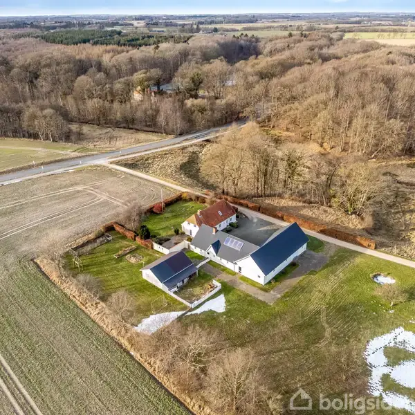 Et huskompleks med flere bygninger ligger i en åben natur omgivet af marker og skov. En vej løber ved siden af ejendommen, der har snepletter i haven.