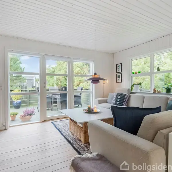 En lys stue med en hjørnesofa og sofabord. Store vinduer og glasdør åbner ud til en terrasse med planter. Væggene er hvide, hvilket skaber en luftig atmosfære.