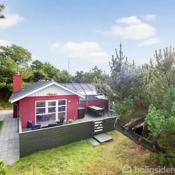 Huset er rødt med gråt tag, har store vinduer og ligger på en terrasse omgivet af træer. Skiltet "Patina" er monteret over terrassen. Skyfri blå himmel.