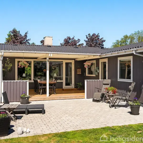Terrasse med sorte loungemøbler står på flisebelagt areal foran et gråmalet hus med store vinduer. Blomsterkrukker og grønne træer omgiver området og skaber en hyggelig atmosfære.