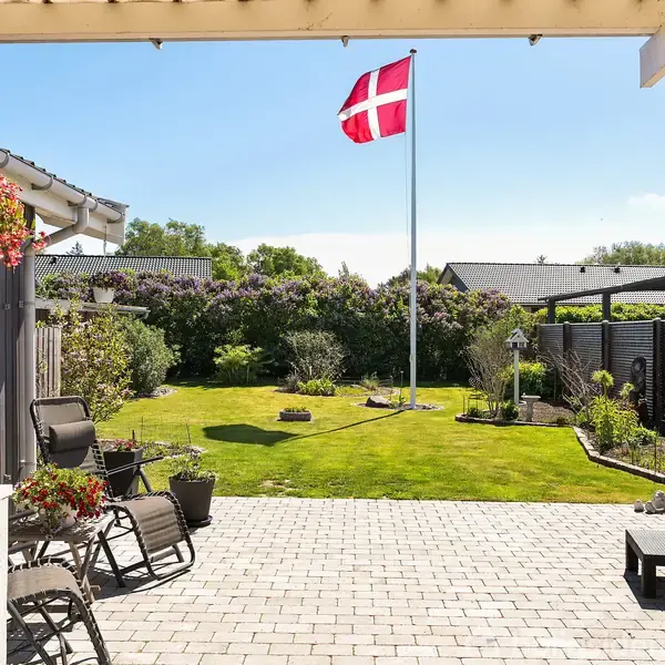 Flagstang med Dannebrog vajer i en solrig have omgivet af grønne buske og hegn. En terrasse med fliser, blomsterkrukker og havemøbler danner forgrunden.