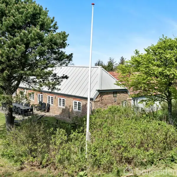 Et murstenshus med gråt tag står midt i en grøn have. En flagstang uden flag står foran huset, omgivet af træer og buske under en klar blå himmel.
