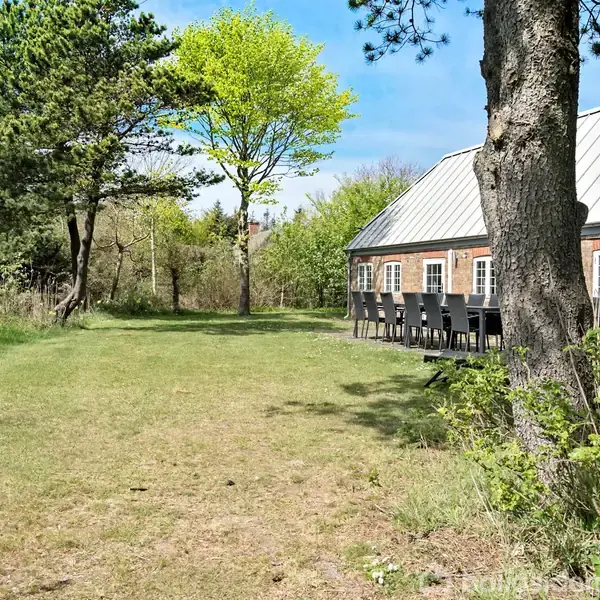 En lang, græsbevokset have strækker sig foran et bagvedliggende murstenshus med et lyst tag. Langs husets facade står en række sorte havestole. Omkring er der grønne træer og buske.