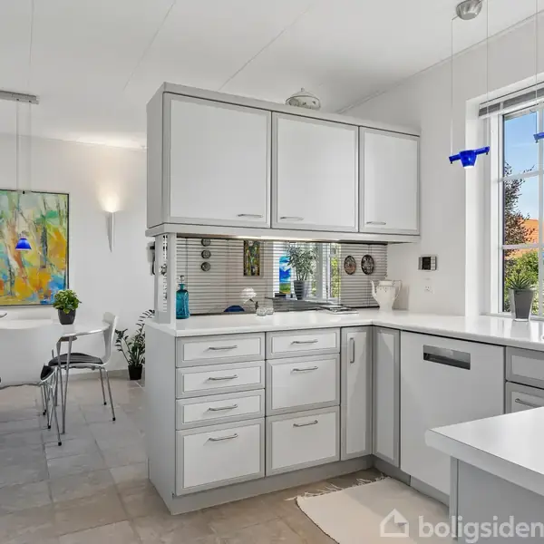 Køkken med hvide skabe og skuffer står i moderne stil, set mod spisestue med abstrakt maleri. Blå lamper hænger over spisebordet i rummeligt, lyst interiør.