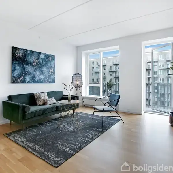 Grøn sofa står i stue ved siden af et lille bord og stol. Kunstværk hænger på væggen. Store vinduer viser udsigt over moderne lejlighedsbygning. Lys og rummelig indretning.