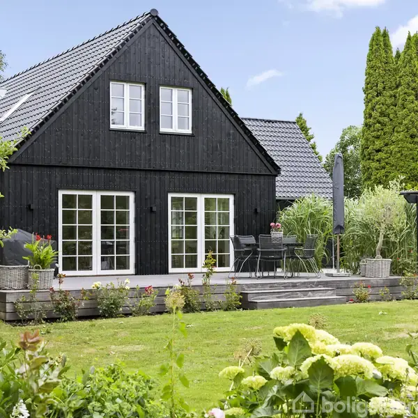 Sort træhus med hvide vinduer, omgivet af frodig have med blomster og buske. På terrassen står et bord og stole. Bagved er der høje træer og en klar blå himmel.