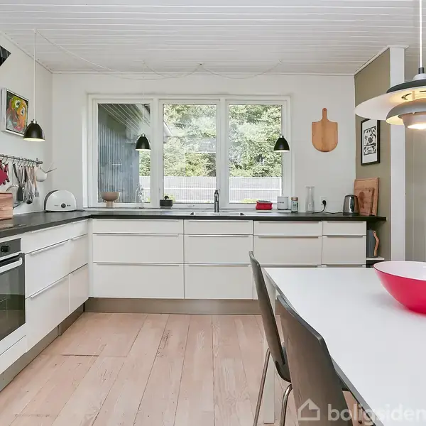 Et moderne køkken med hvidt interiør og træbord udfører daglige madlavningsaktiviteter i lyse omgivelser. En rød skål på bordet tilføjer farve. Vinduer afslører grønne omgivelser udenfor.