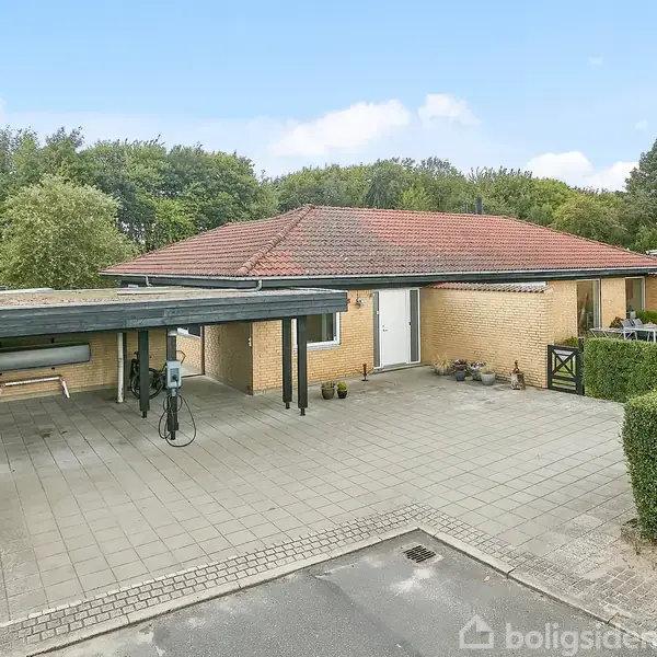 Et gult murstenshus med rødt tag står ved siden af en carport med overdækning. Flisebelagt indkørsel og omkranset af grønne hække. Skovområde i baggrunden.