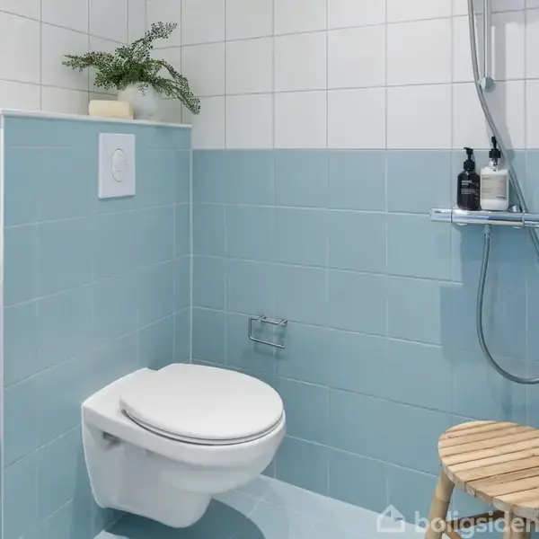 Et badeværelse med et væghængt toilet ved siden af en brusekabine. Blå og hvide fliser dominerer væggene. Der står en skammel med træfinish og en hylde med sæbe.