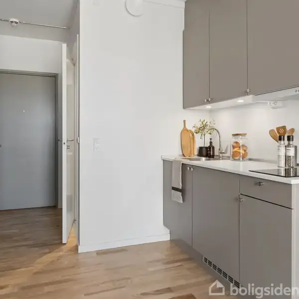 Et lille køkken med grå skabe og et komfur. Bordet har køkkenredskaber og planter. I baggrunden ses en åbentstående dør til en entré. Gulvet er af træ.