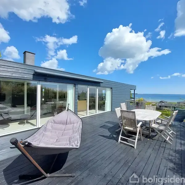 Et moderne hus med store vinduer har en terrasse med en hængestol og et rundt spisebord med stole. Terrassen har udsigt over havet under en klar blå himmel.