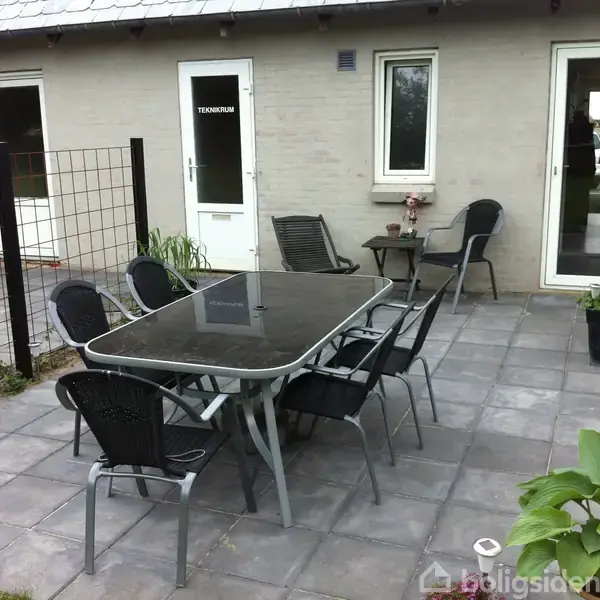 Terrassebord med seks sorte stole står på flisebelagt terrasse. En dør markeret med "Teknikrum" ses i baggrunden, omgivet af lyse murstensvægge, et lille bord og planter.