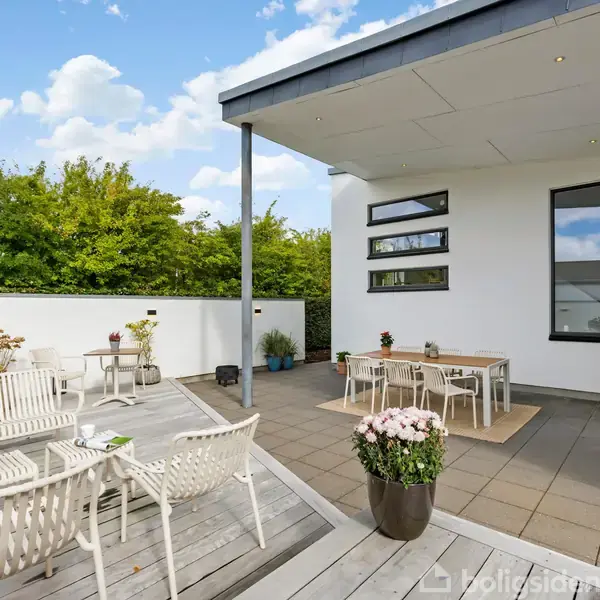 Terrasse med hvide havemøbler står stille på lyse træplanker. I kontekst er der en moderne hvid murstensbygning med et fladt tag og store vinduer, omgivet af grønne træer.