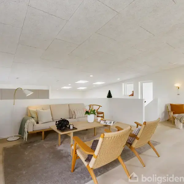 Stue med sofa og to lænestole omkring et sofabord. Gulvlampe ved siden af sofaen. Rummet har lyse vægge, store vinduer og moderne indretning. Indretningen skaber en lys og rummelig atmosfære.