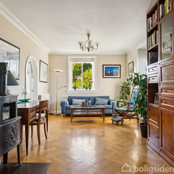 Brændeovn står i et lyst stueområde ved siden af et lille træbord. Sofa og planter placeres nær vinduet. Reoler fyldt med bøger og dekorationer fylder højre side.