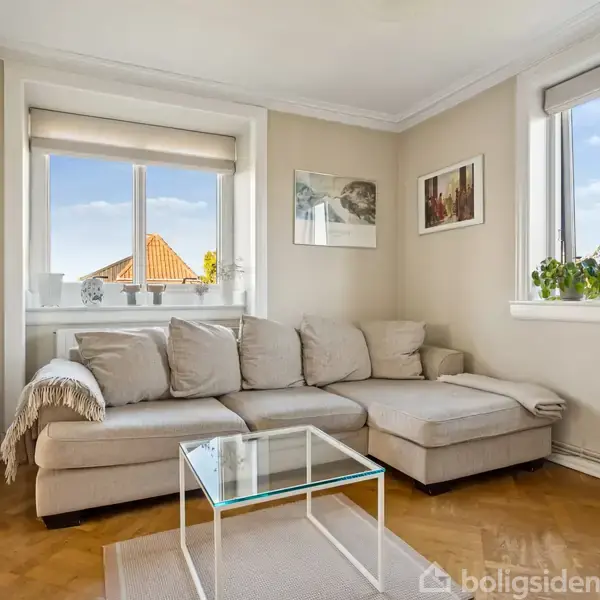 Beige hjørnesofa med puder står i en lys stue ved vinduer. Glasbord foran sofaen. Billeder hænger på væggene, og planter er placeret på vindueskarmen.