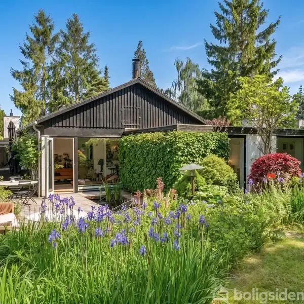 Et hus dækket af grøn beplantning ligger i en frodig have fyldt med blomster og træer. Døre er åbne mod en terrrasse, der inviterer til udendørs afslapning.