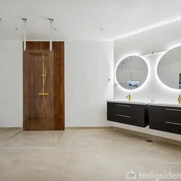 Badeværelse med stor, glasindkapslet bruser med træpaneler og guldarmatur; to ovale spejle over sorte dobbeltvaskeskabe; rumlys og håndklæder i reol mod hvid væg.