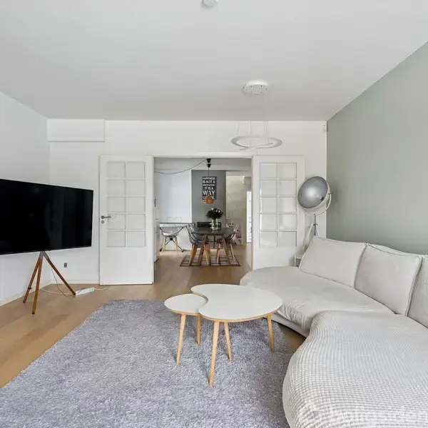 En lys stue med en stor, lys beige sofa, der står langs højre væg. Et rundt sofabord står på et gråt tæppe. Et fladskærms-TV er på venstre væg. I baggrunden ses en spisestue.