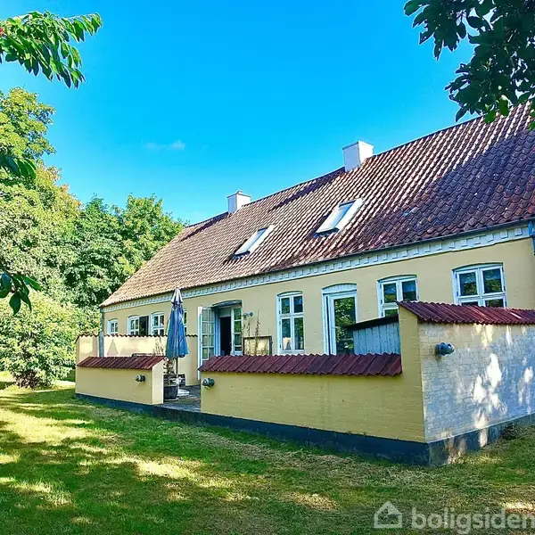 Et gult, klassisk hus med rødt tegltag står i en solrig have med grønne træer. Terrasse med parasol anes langs huset, omgivet af frodig natur.