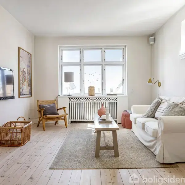 Sofa står stille, placeret i et lyst stueområde. Ved siden af er der et lille bord og en stol. Vinduerne i baggrunden lader naturligt lys strømme ind. En kurv og fjernsyn hænger på væggen.