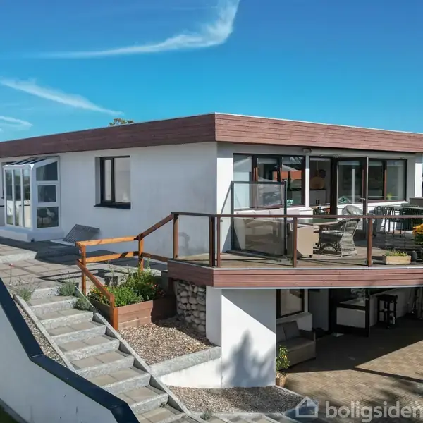 Et moderne hus står på en skråning med en stor terrasse med glasrækværk. Terrassen er indrettet med bord og stole. Huset har hvid facade og fladt tag.
