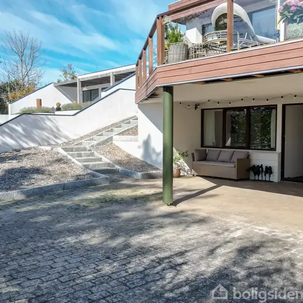 Terrasse med møbler står stille under en overdækning forbundet med et moderne hus. Omgivelserne inkluderer en stentrappe og grønne træer i baggrunden.